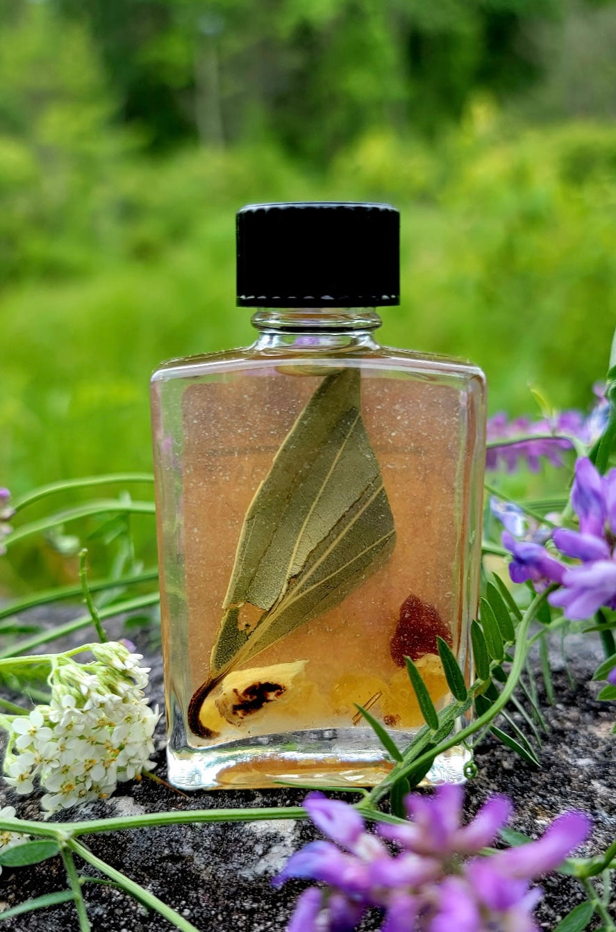 Small bottle with a black cap containing a leaf and other elements, surrounded by greenery and flowers.