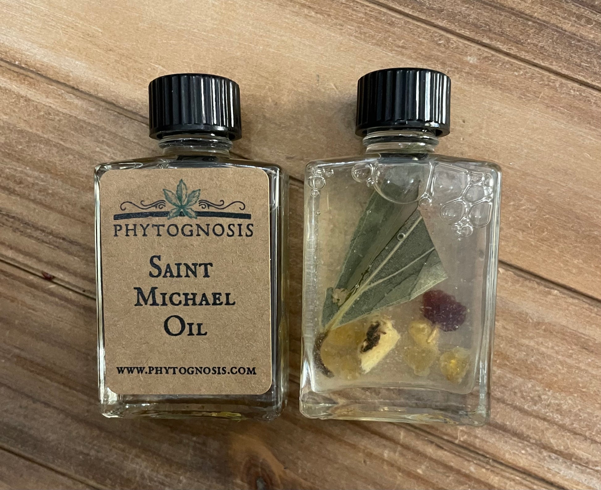 Two small glass bottles with black lids on a wooden surface, one labeled 'Phytognosis Saint Michael Oil'.