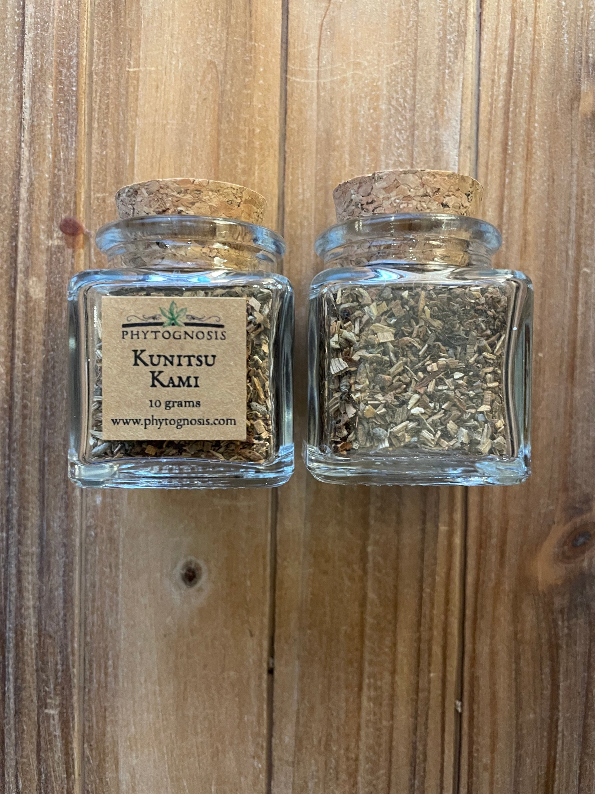 Two small clear glass jars with cork stoppers, filled with dried green herbal or tea-like material, placed on a wooden surface. The lower jar has a paper label reading 'Pittfordnoe Kuntiyo Kami 10 grams'.