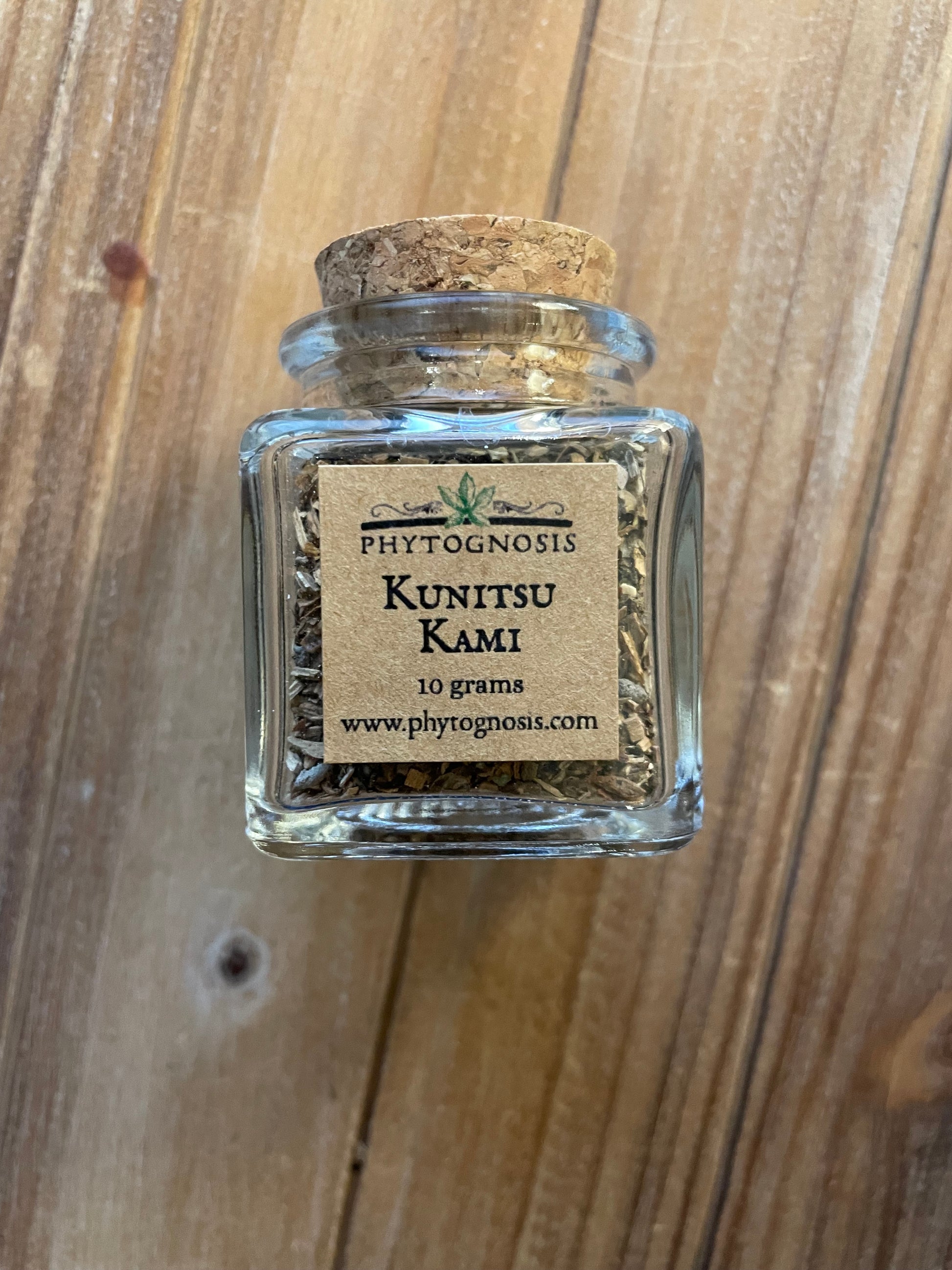 A small clear glass bottle with a cork stopper containing dried plant material, with a tan paper label reading 'PHYTOGNOESIS KUNITSU KAMI 10 grams' and a website, placed on a wooden surface.