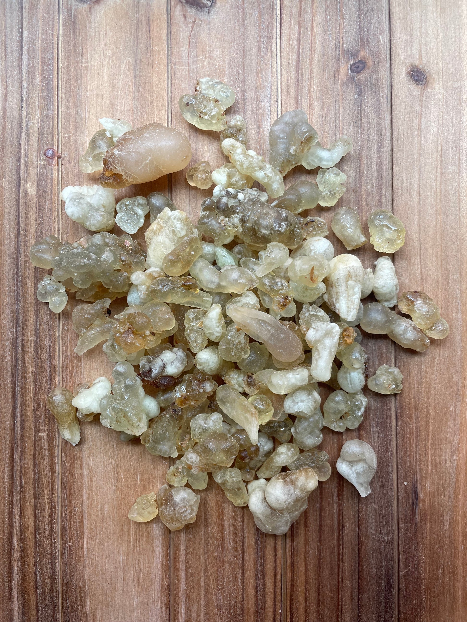 A pile of translucent, gelatinous, off-white and pale yellow dried botanical pieces, frankincense resin pieces, arranged on a brown wooden surface.