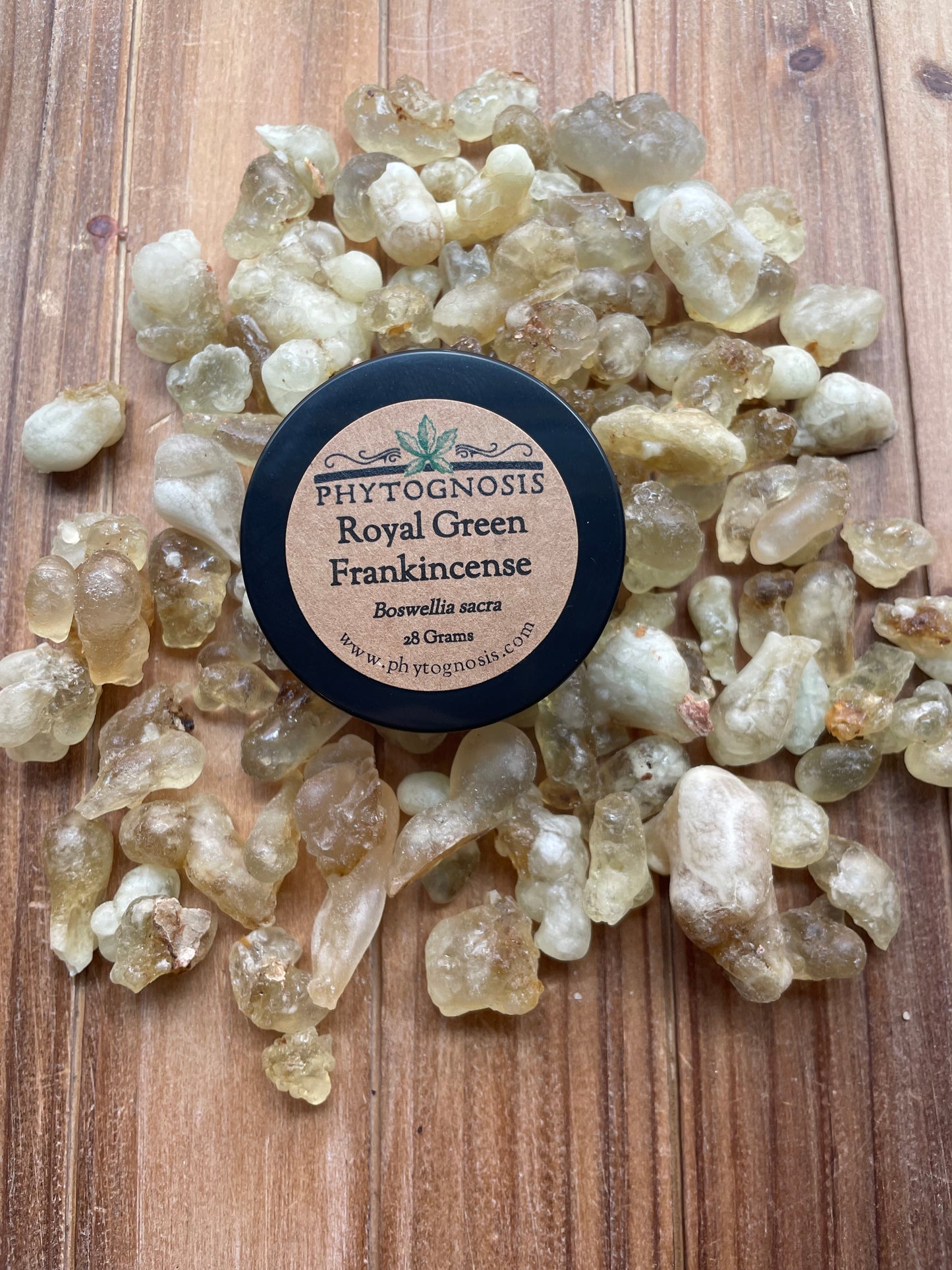 A small round black-lidded tin labeled 'PHYTOGNOSIS Royal Green Frankincense' sits on a wooden surface surrounded by translucent raw frankincense resin pieces.