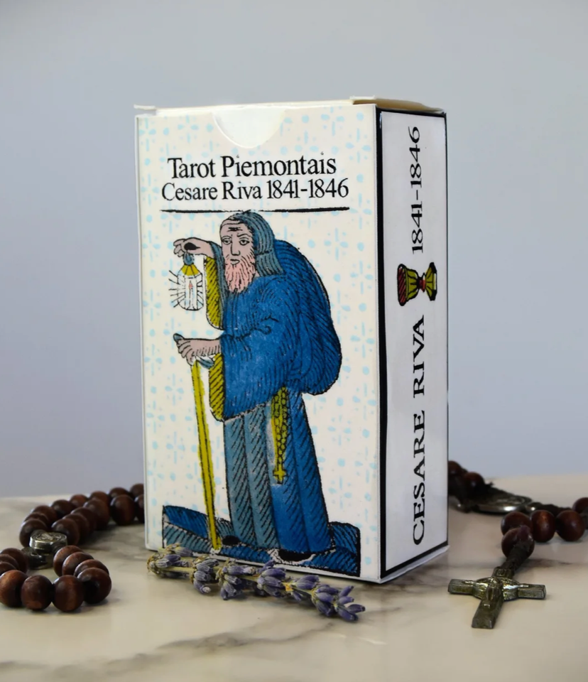 A boxed tarot card deck labeled 'Tarot Piemontais Cesare Riva 1841-1846' stands on a marble surface with rosary beads and a cross around it. The box shows an illustrated figure in blue robes holding a staff, with a pale blue patterned background.